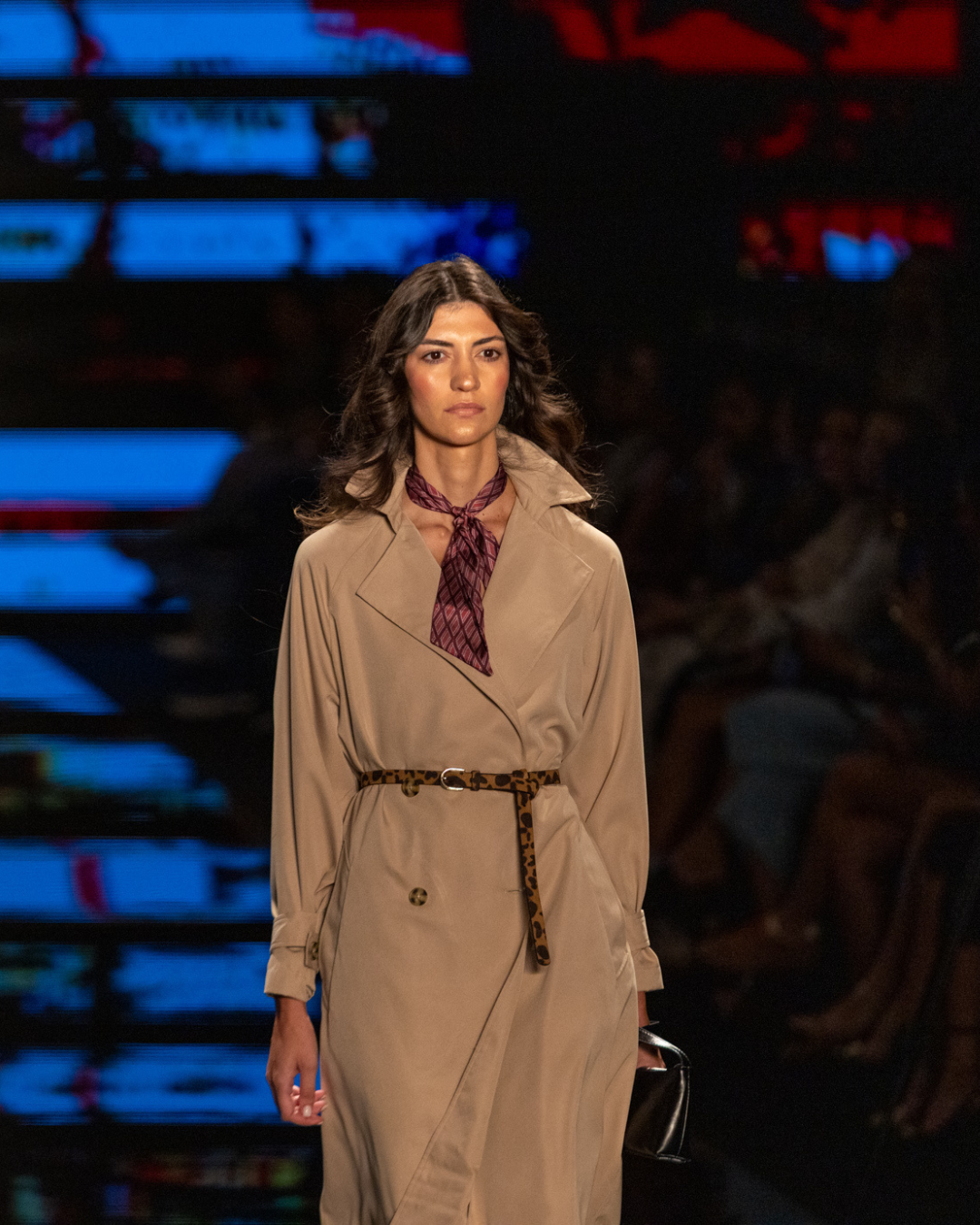 Acessórios tendência do inverno 2026: modelo desfila com lenço vinho e cinto animal print sobre trench bege, clima