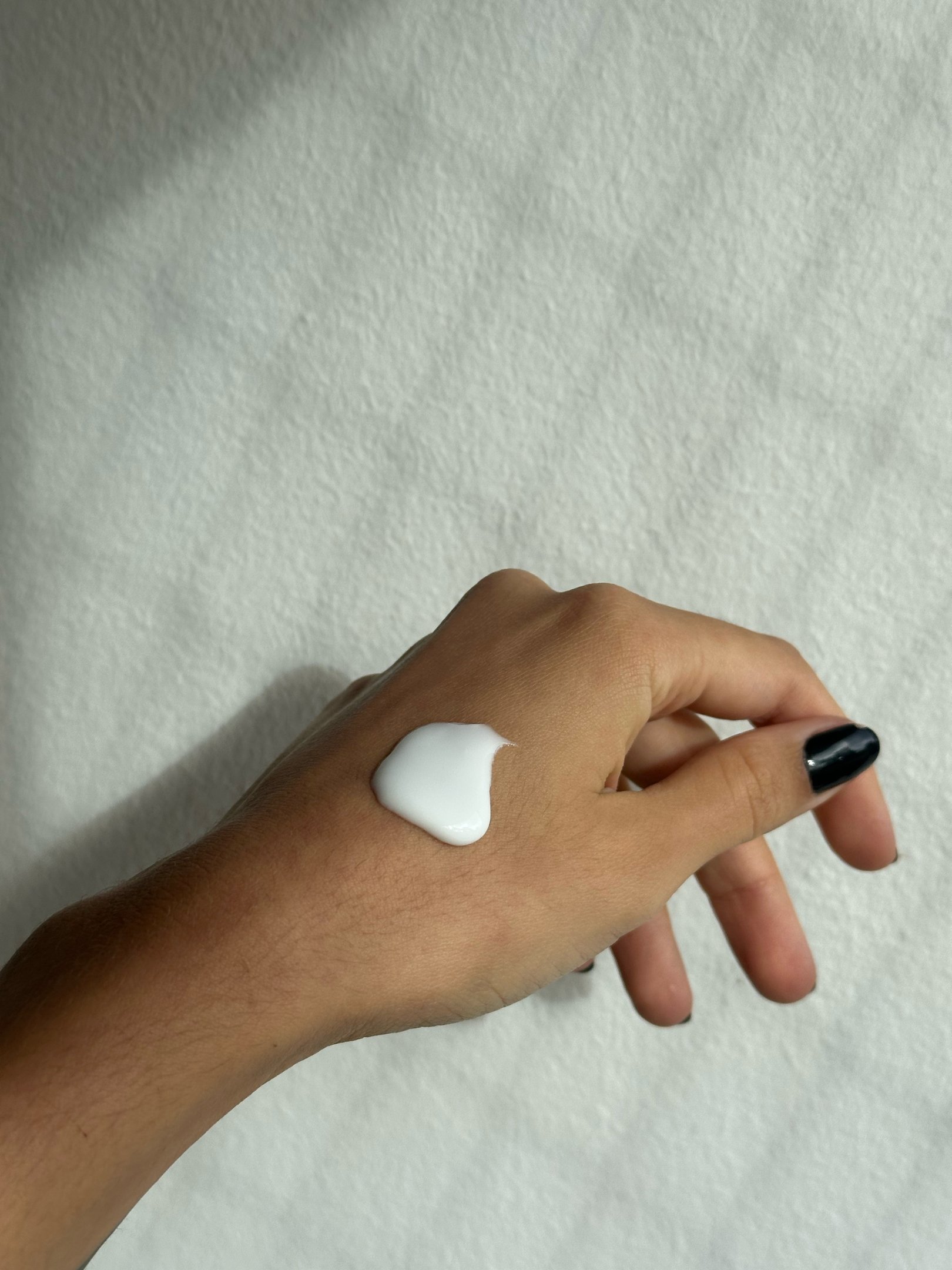 Loção corporal da Moroccanoil sobre mão em pose suave, creme branco e unha preta contrastam com fundo claro minimalista