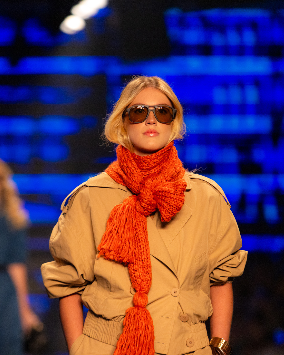 Acessórios tendência do inverno 2026: modelo posa com óculos oversized e cachecol laranja em tricô, look bege imponente.