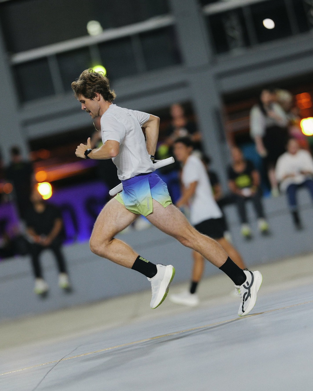 Corre 5 em ação: corredor salta com camiseta branca, short degradê e tênis branco em clima urbano vibrante.