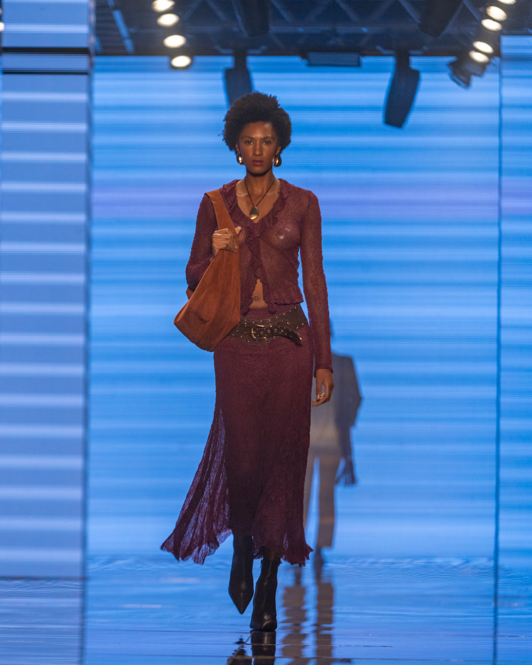 Acessórios tendência do inverno 2026: modelo posa com bolsa hobo, cinto largo e botas pretas em look vinho sofisticado.