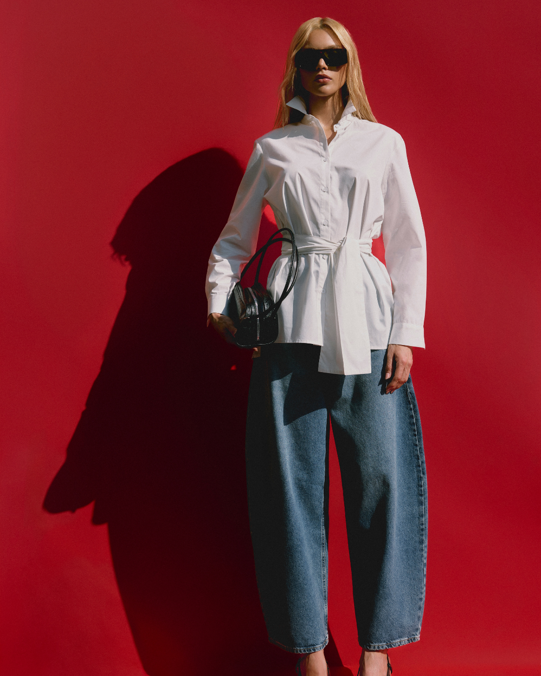 Como usar jeans de forma estilosa: modelo posa com camisa branca acinturada, jeans amplo azul e bolsa preta em fundo