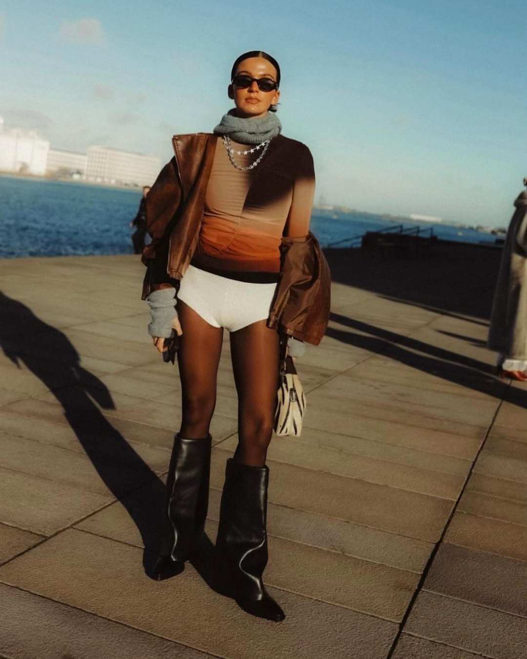 Modelo posa com bota capa preta, body degradê, jaqueta marrom e gola cinza à beira-mar em clima urbano sofisticado