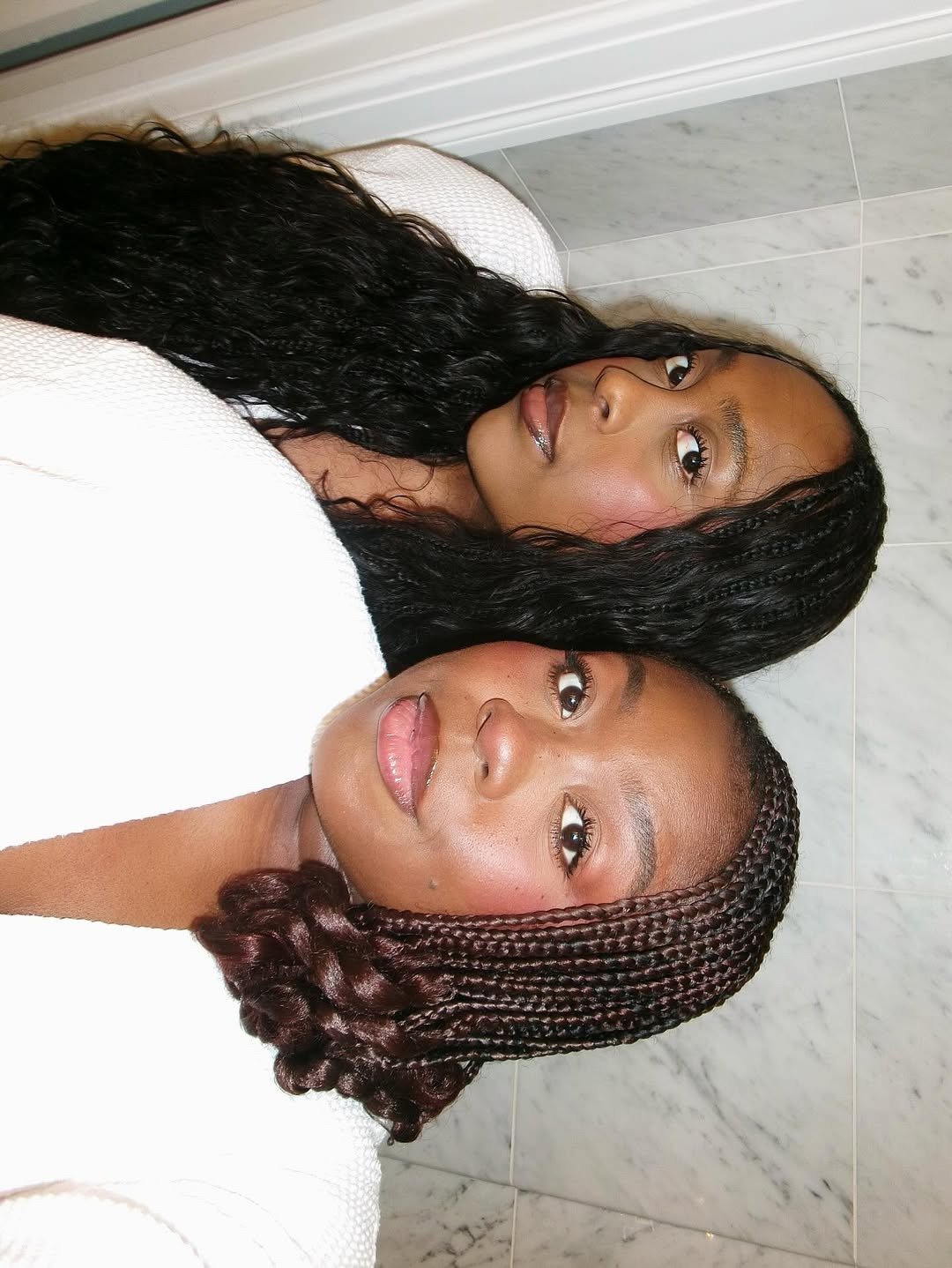 Amigas posam juntas com tricô branco, tranças e cachos escuros em close elegante diante de parede marmorizada clara