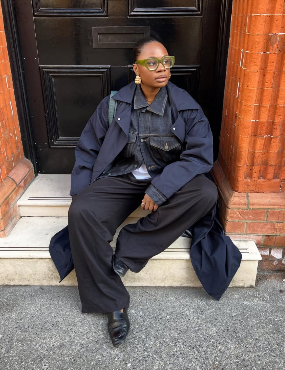 Modelo posa sentada com armações estilosas para óculos de grau verdes, trench azul-marinho e jeans escuro diante de porta.