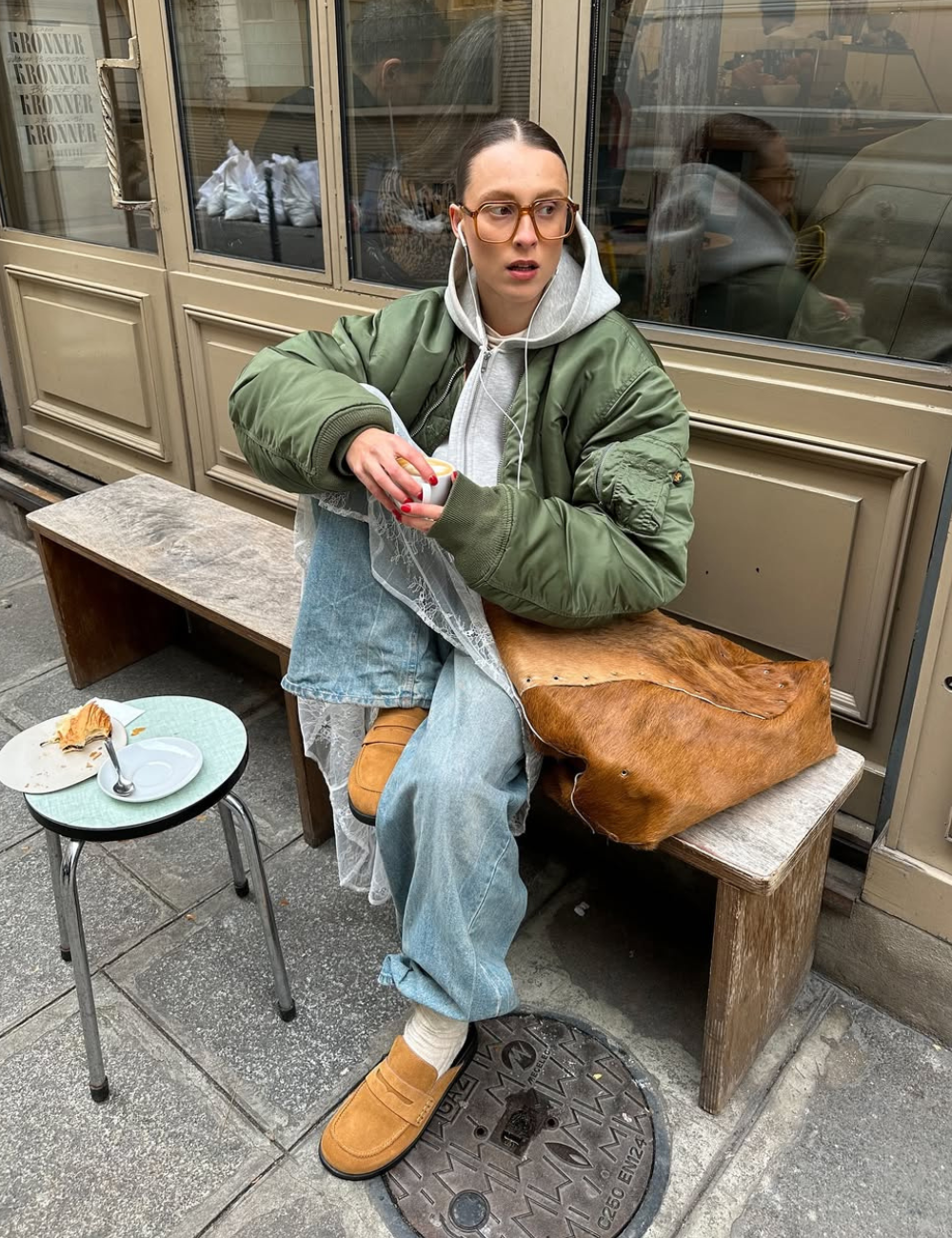 Modelo posa com armações estilosas para óculos de grau, bomber verde, moletom cinza e jeans amplo em clima urbano casual.