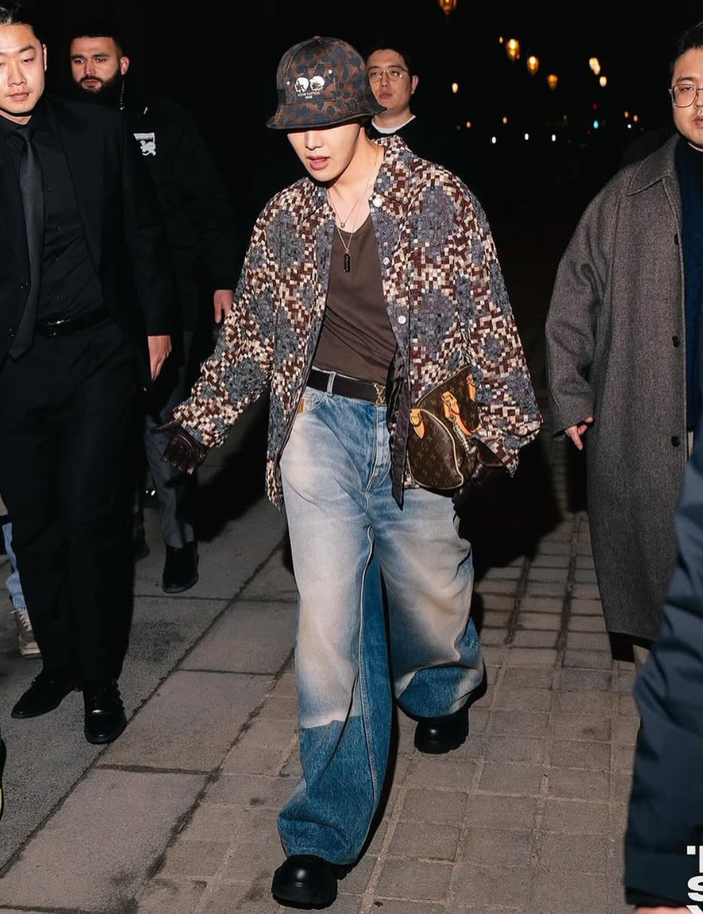 BTS caminha à noite com jaqueta texturizada, jeans amplos degradê, boné e bolsa marrom em clima urbano confiante