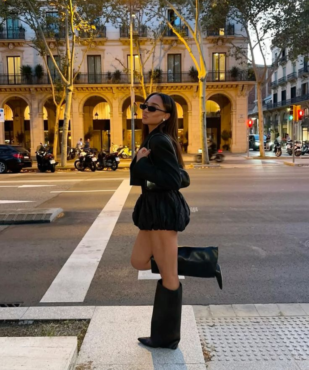 Modelo posa na faixa com look preto bufante e bota capa de couro, óculos escuros e clima urbano sofisticado.