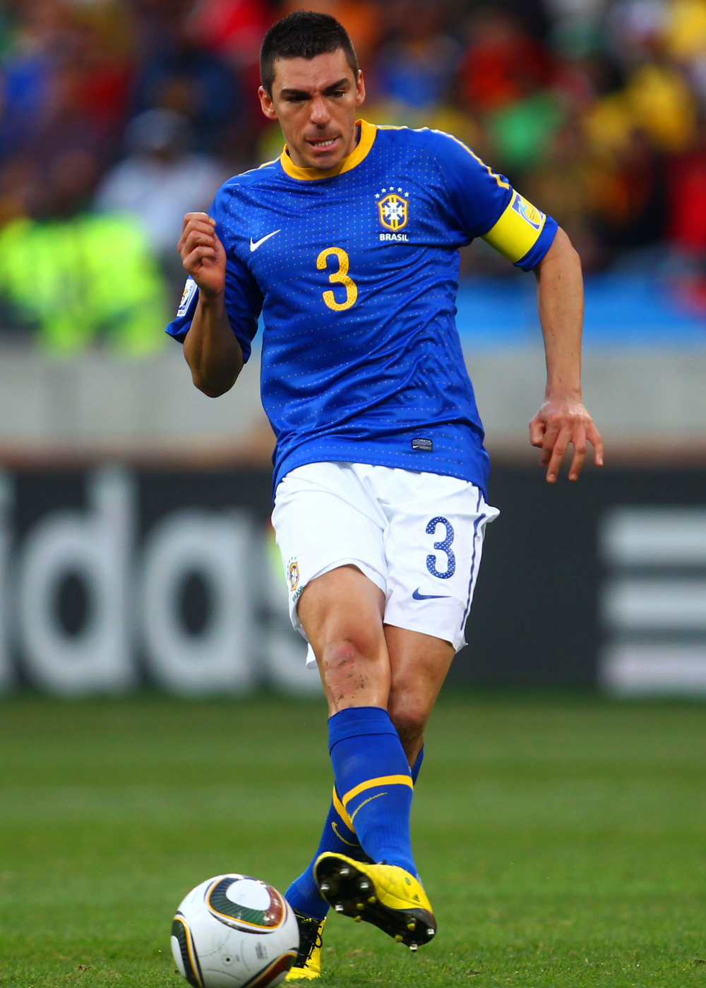 Modelo corre em campo com camisa do Brasil azul de tecido esportivo, short branco e braçadeira amarela.