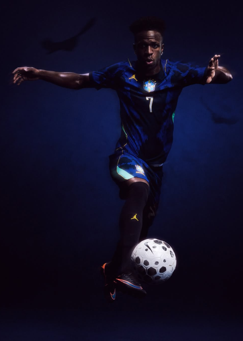 Modelo salta com camisas do Brasil azul-marinho listradas, short esportivo e bola, em clima intenso e dinâmico.