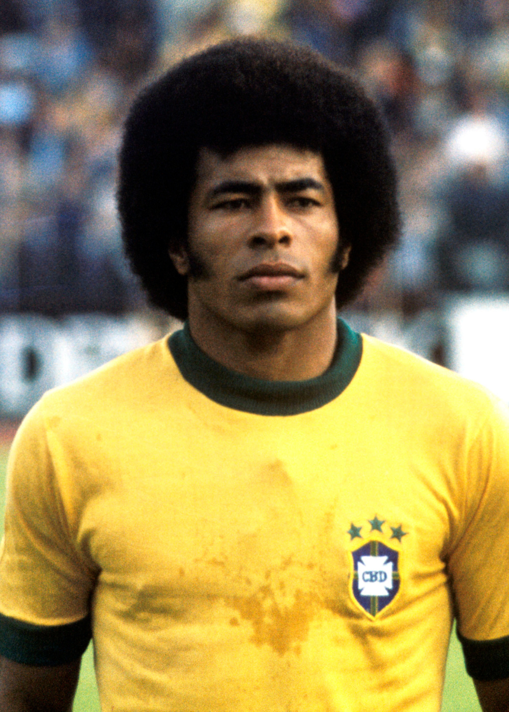 Camisas do Brasil em amarelo e verde, modelo posa sério com uniforme retrô de malha e escudo CBD em campo desfocado.