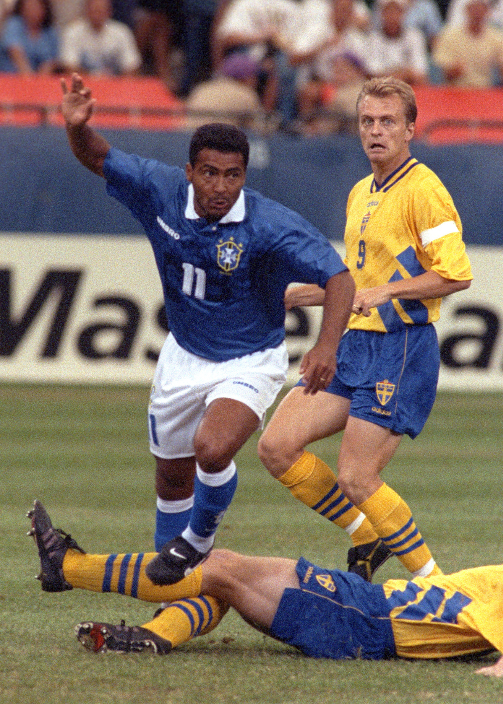 Camisas do Brasil em azul com gola branca e amarelo vibrante marcam jogada tensa entre jogadores em campo lotado.