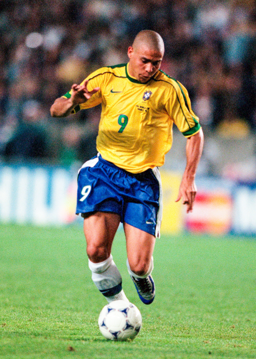Camisas do Brasil em amarelo e verde com short azul; jogador avança com a bola em campo, em clima vibrante.