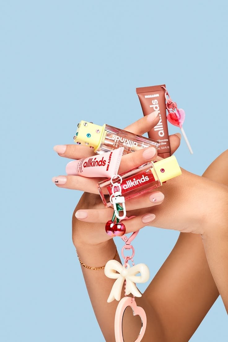 Mãos com unhas nude exibem gloss allkinds rosa e marrom com pingentes fofos em fundo azul. Texto: allkinds spvillio