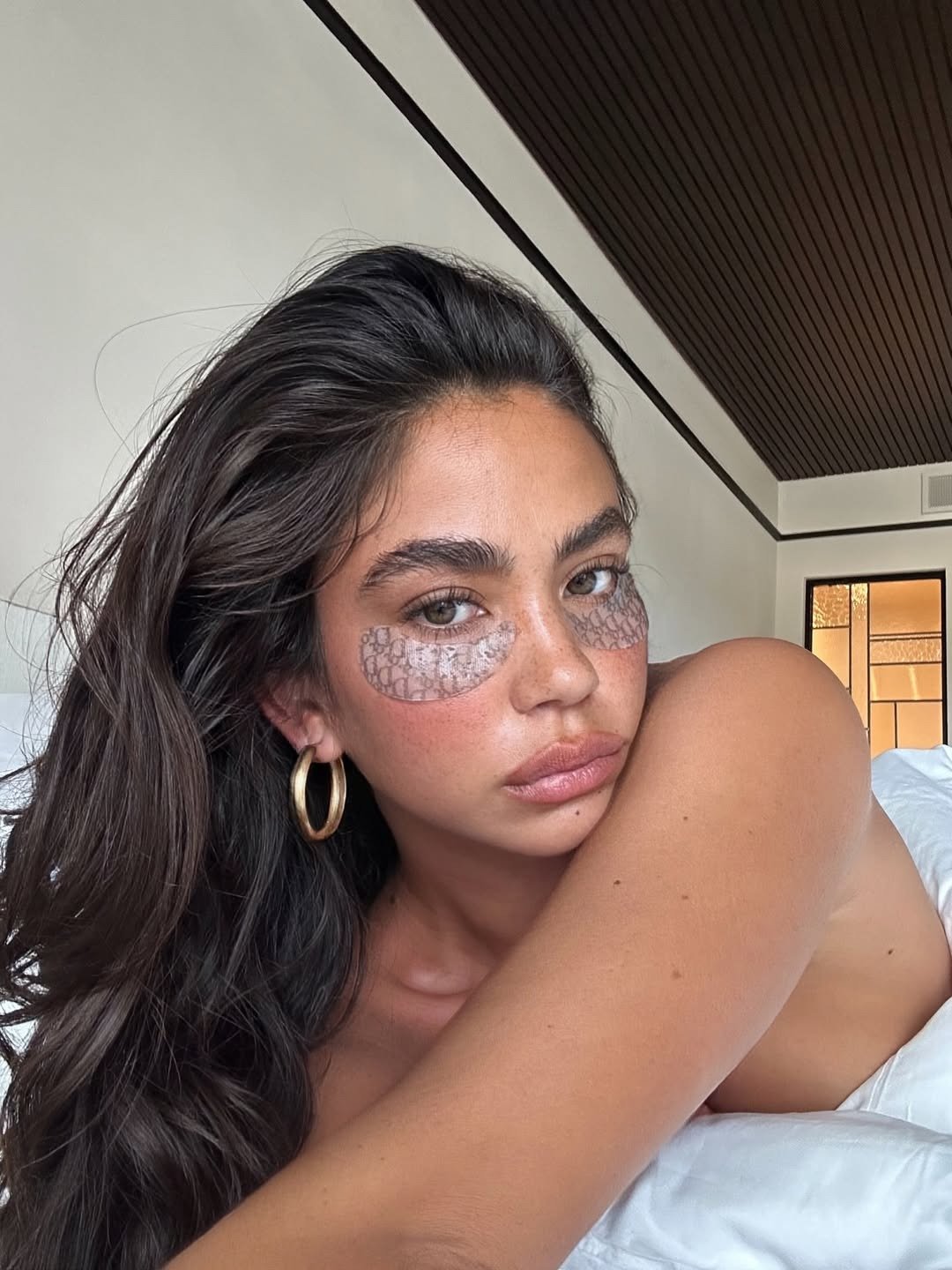 Modelo posa com cremes para olheiras em adesivos sob os olhos, argolas douradas e clima íntimo em tons neutros.