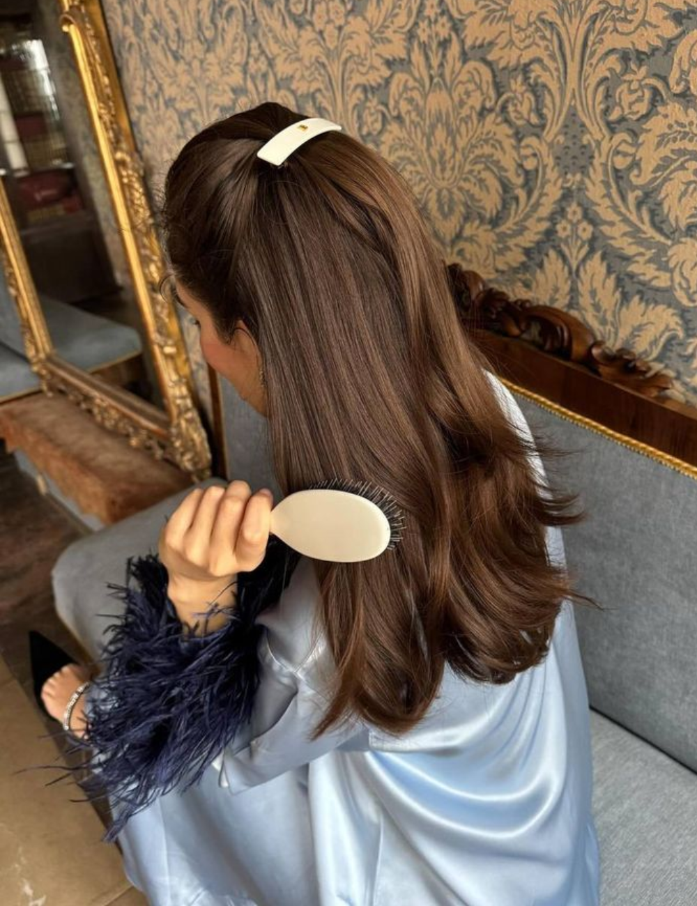 Como acabar com frizz no cabelo liso: mulher sentada escova fios castanhos lisos com robe acetinado azul-claro.