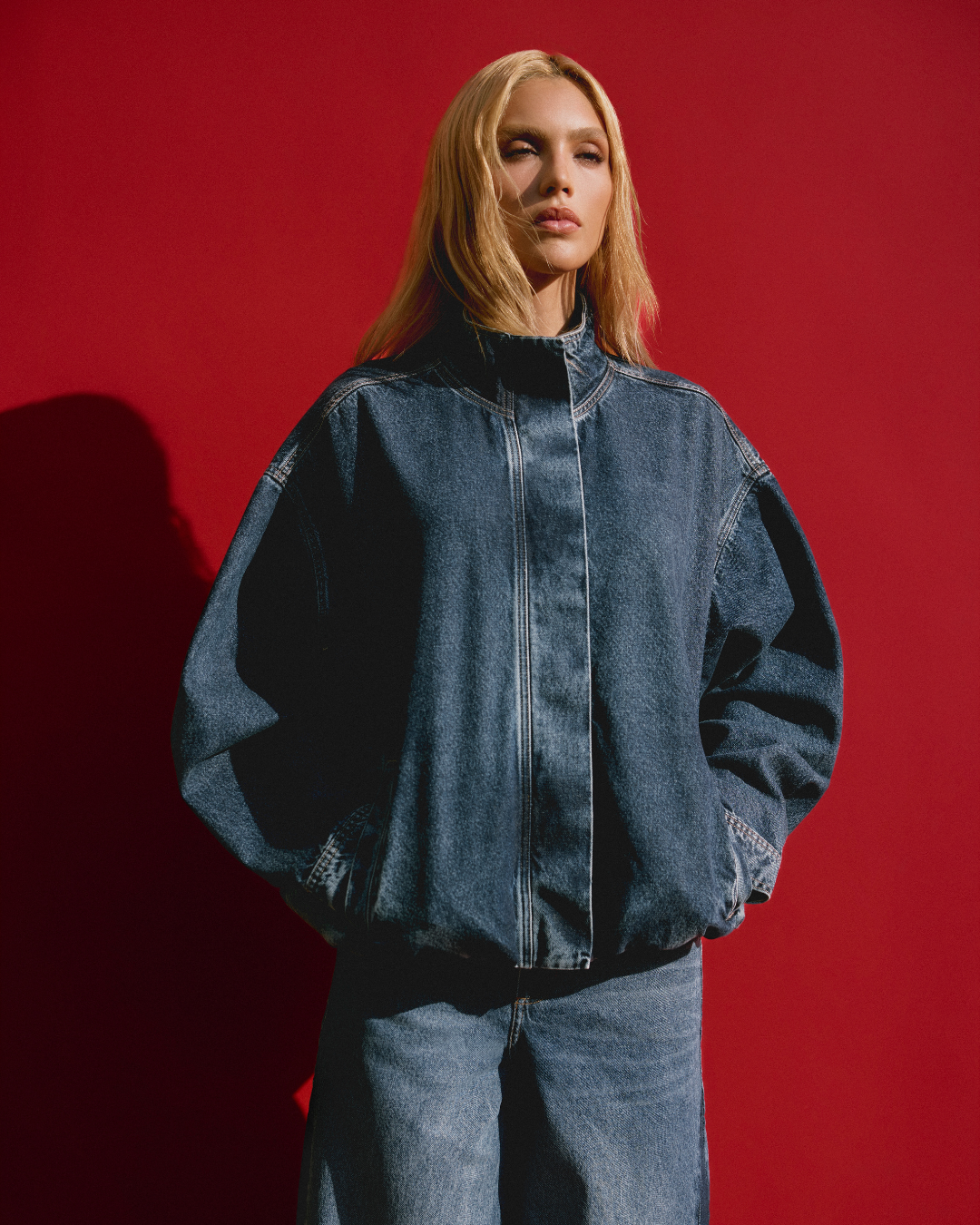 Como usar jeans de forma estilosa: modelo posa com jaqueta oversized e calça reta azul contra fundo vermelho marcante.