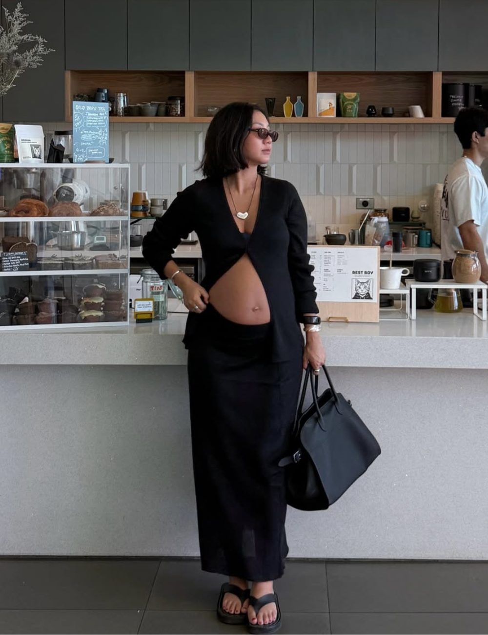 Look all black com cardigan recortado e saia longa; modelo posa no café com bolsa maxi, óculos escuros e elegância urbana.