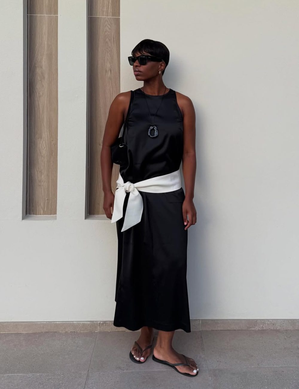 Modelo posa em look all black acetinado com regata e saia midi, faixa branca na cintura e atitude elegante.