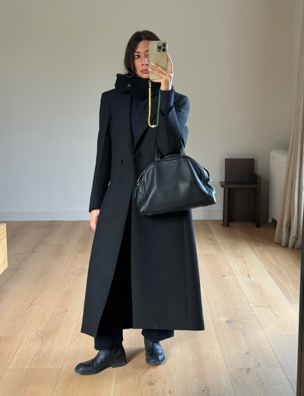 Look all black com modelo posa de sobretudo reto, tricô e botas de couro, bolsa estruturada em ambiente minimalista sóbrio.