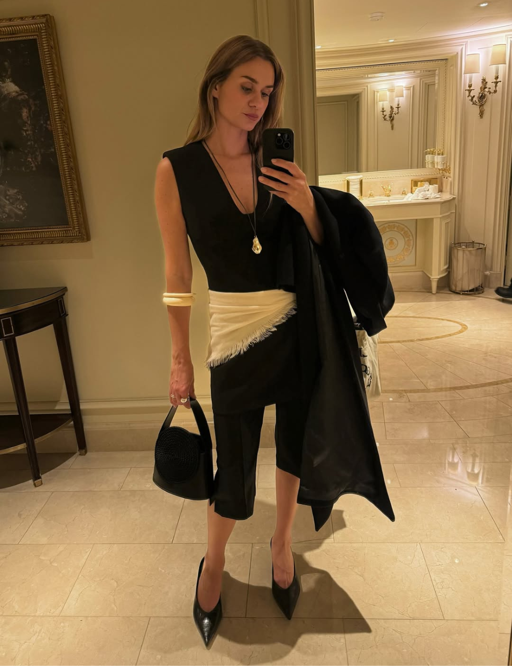 Modelo posa com look all black de decote V e saia midi assimétrica, faixa branca e elegância sofisticada no espelho.