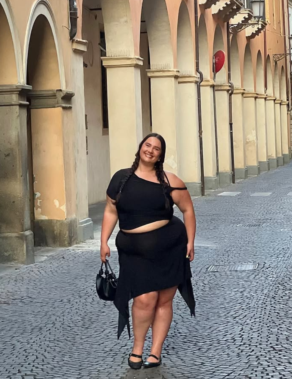 Modelo posa com look all black de top assimétrico e saia drapeada, em rua de arcos bege, com elegância leve.