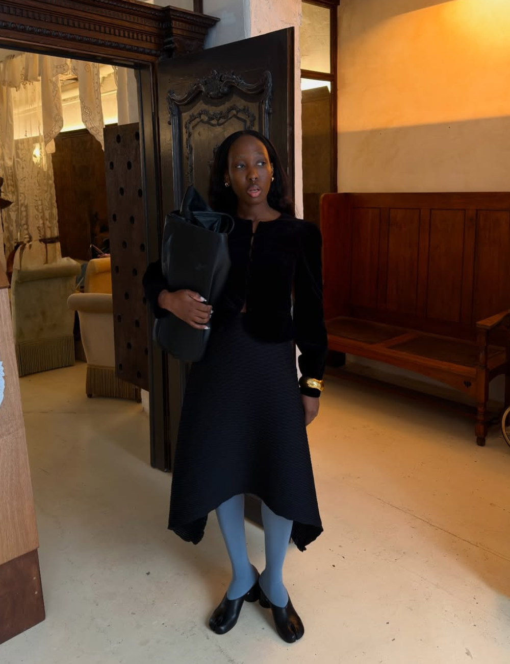 Modelo posa com look all black de veludo e saia midi texturizada, bolsa maxi e clima elegante em interior clássico.