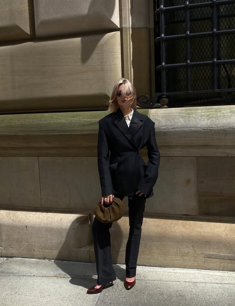 Look all black com blazer acinturado e calça reta; modelo posa ao sol com bolsa marrom diante de fachada clássica.