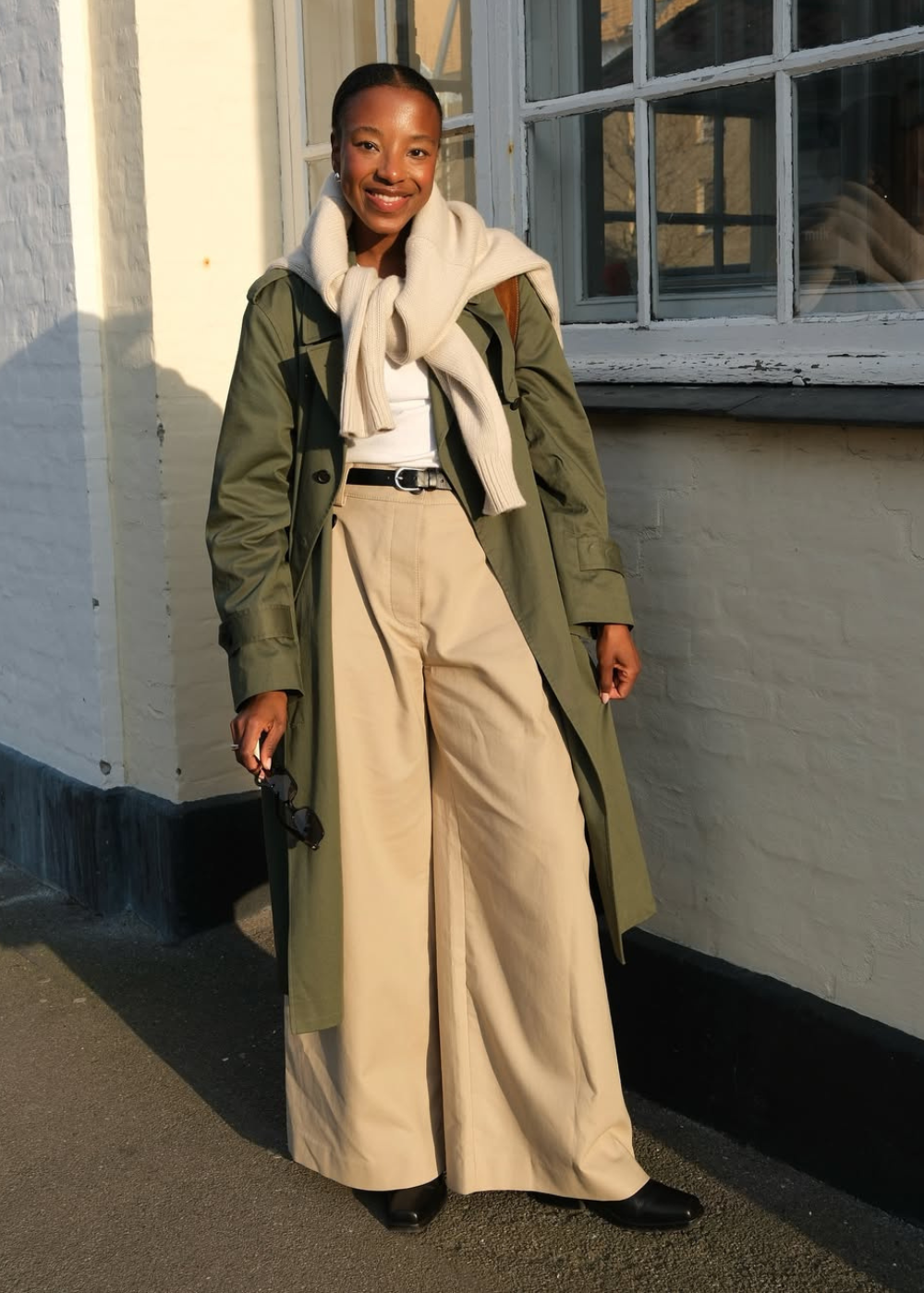 Looks de inverno 2026: modelo posa sorrindo com trench verde, tricô creme e pantalona bege em clima elegante.