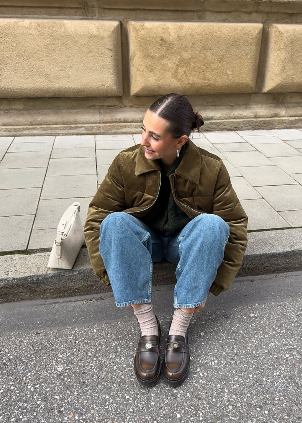 Looks de inverno 2026: modelo posa sentada com jaqueta acolchoada oliva, jeans retos, meias claras e loafers marrons.