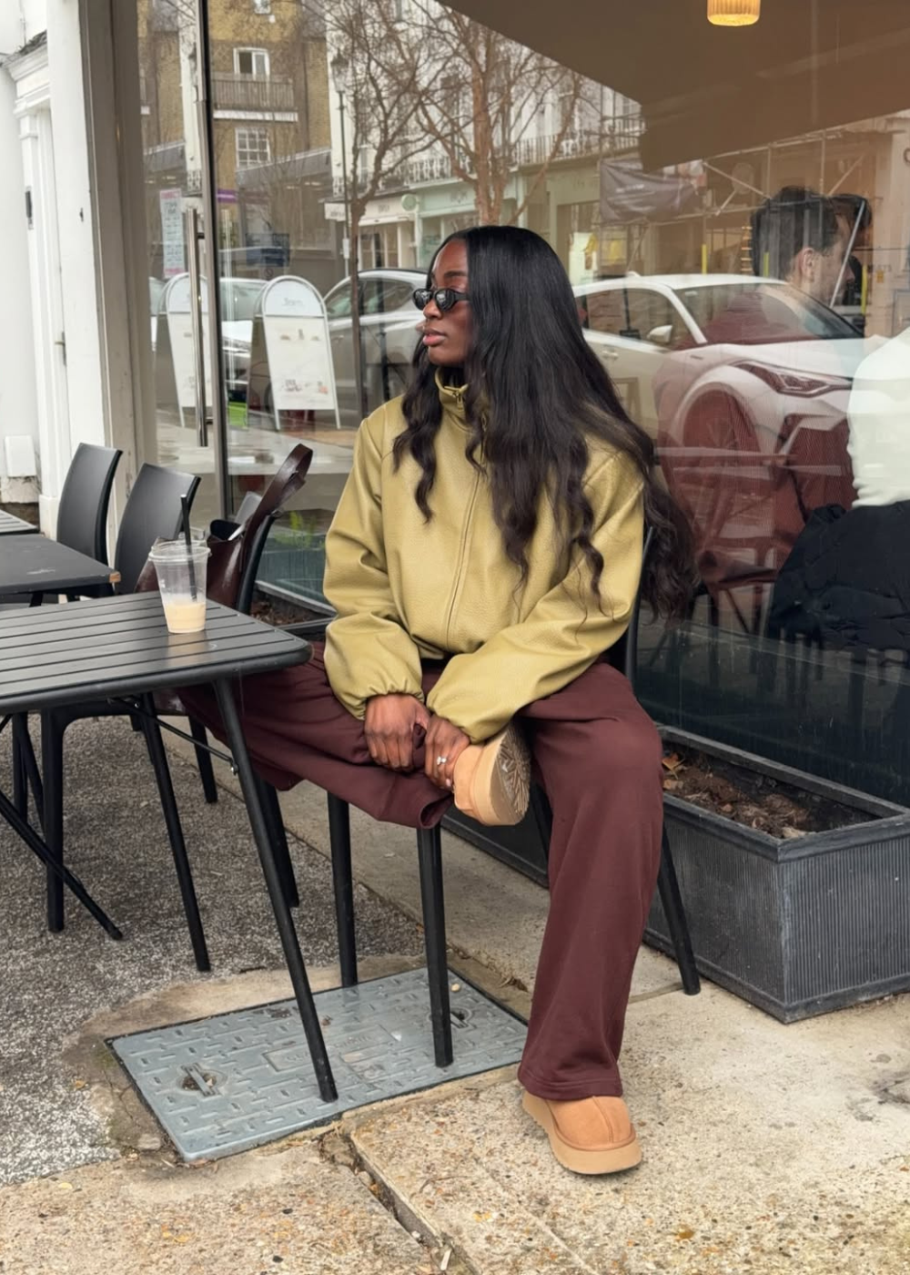 Looks de inverno 2026: modelo posa em jaqueta verde-oliva ampla, calça marrom e botas bege em café urbano acolhedor.