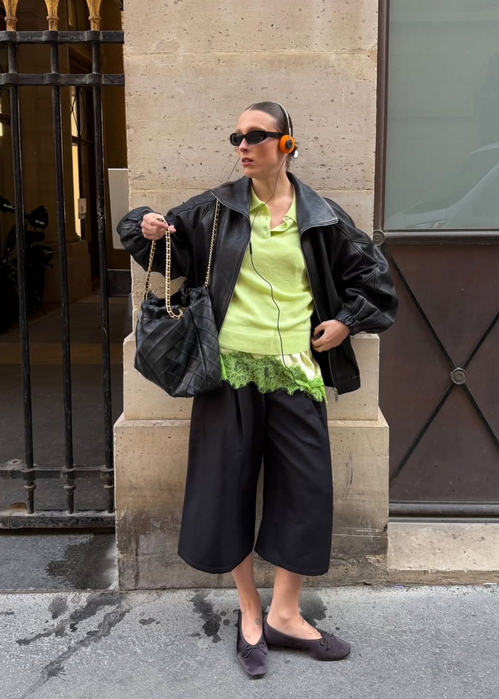 Looks de inverno 2026: modelo posa com jaqueta preta oversized, tricô verde-lima e pantacourt em clima urbano moderno.