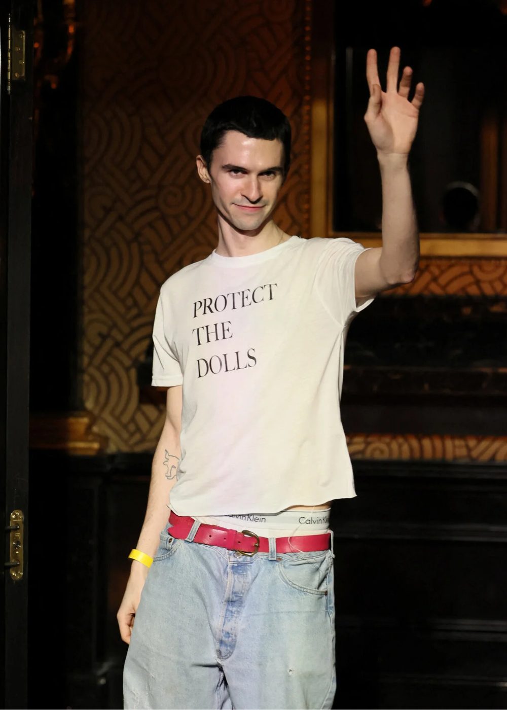 Modelo posa com camisetas branca de algodão, jeans amplo e cinto rosa em clima confiante; Texto: PROTECT THE DOLLS