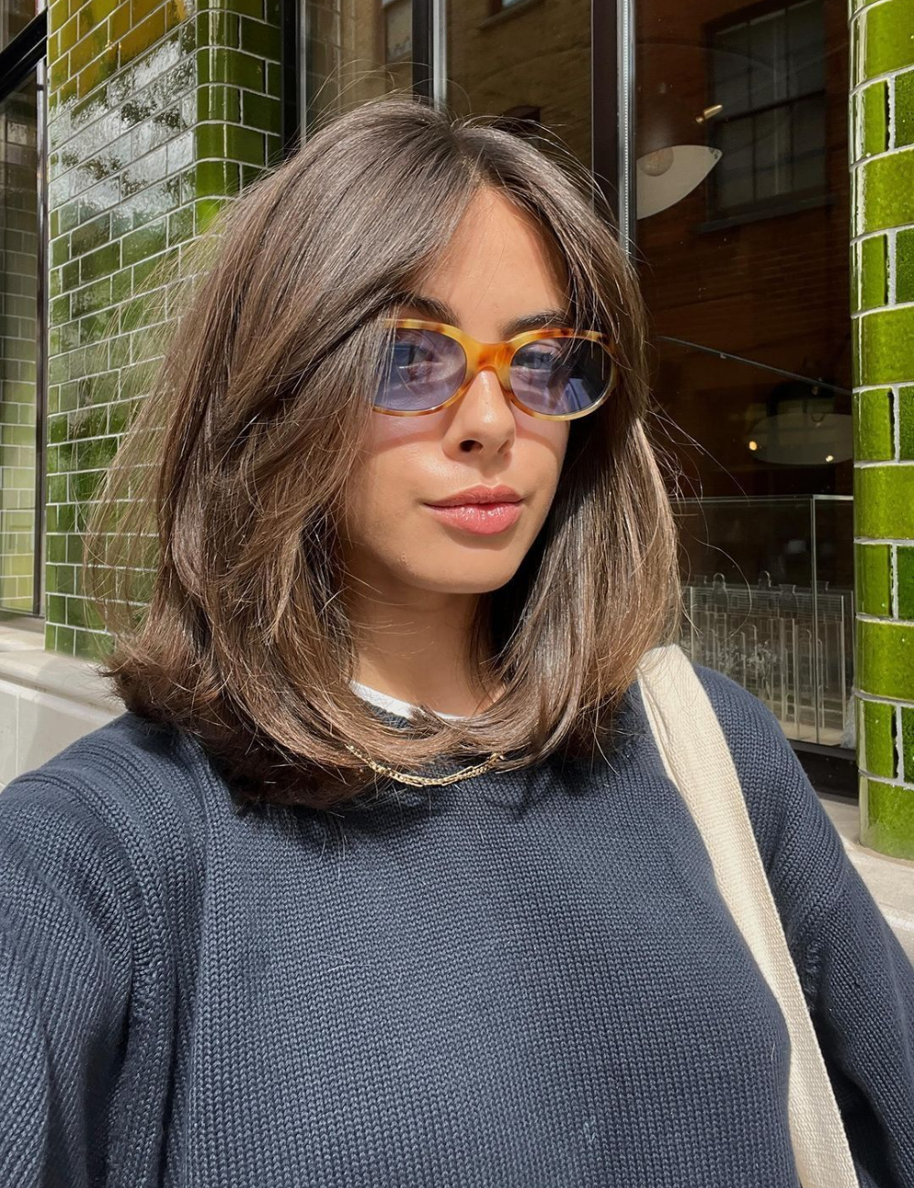 Modelo posa com long bob castanho liso, malha azul-marinho e óculos âmbar diante de azulejos verdes, clima urbano elegante