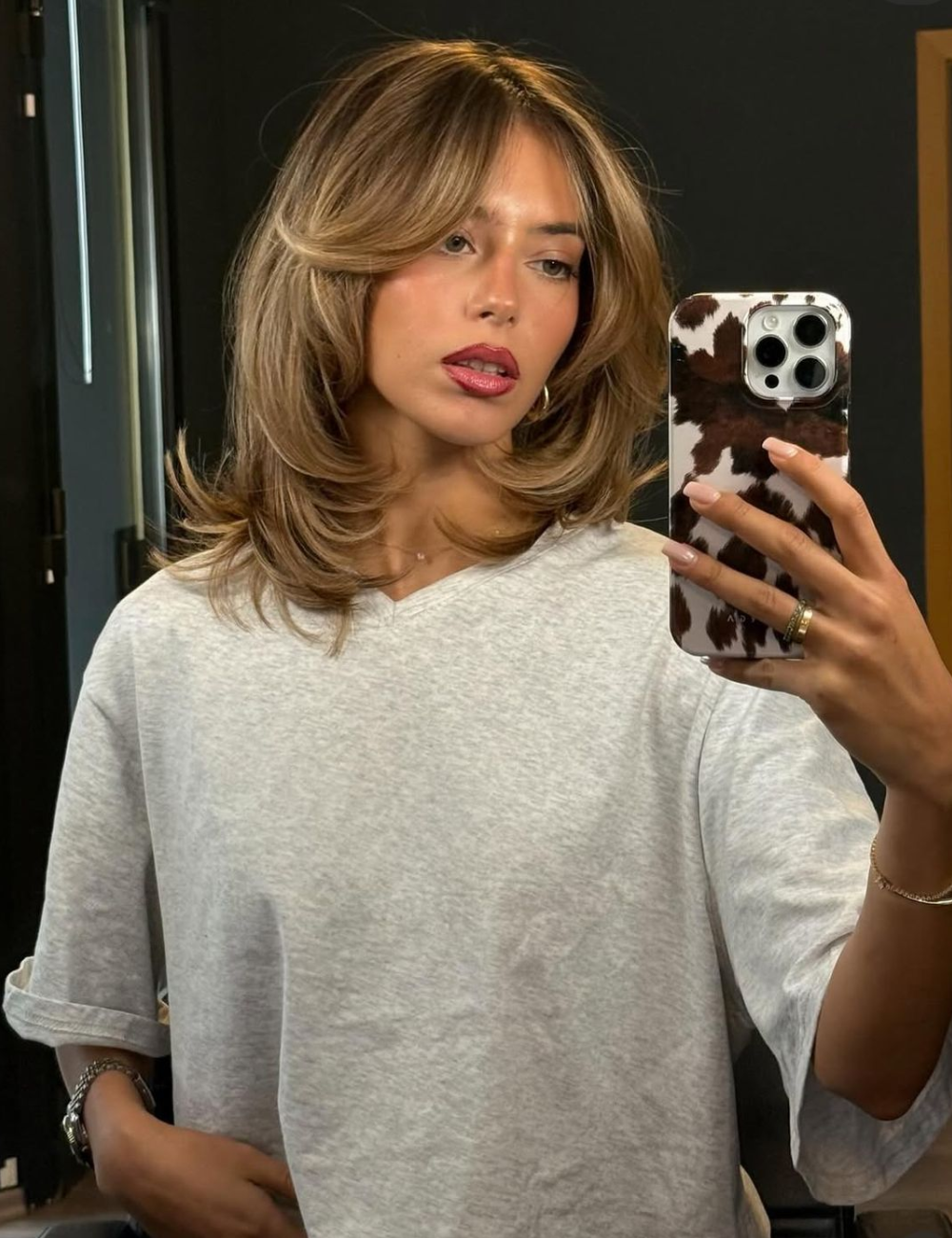 Modelo posa com long bob em camadas loiro mel, camiseta cinza ampla e clima sofisticado em espelho interno.