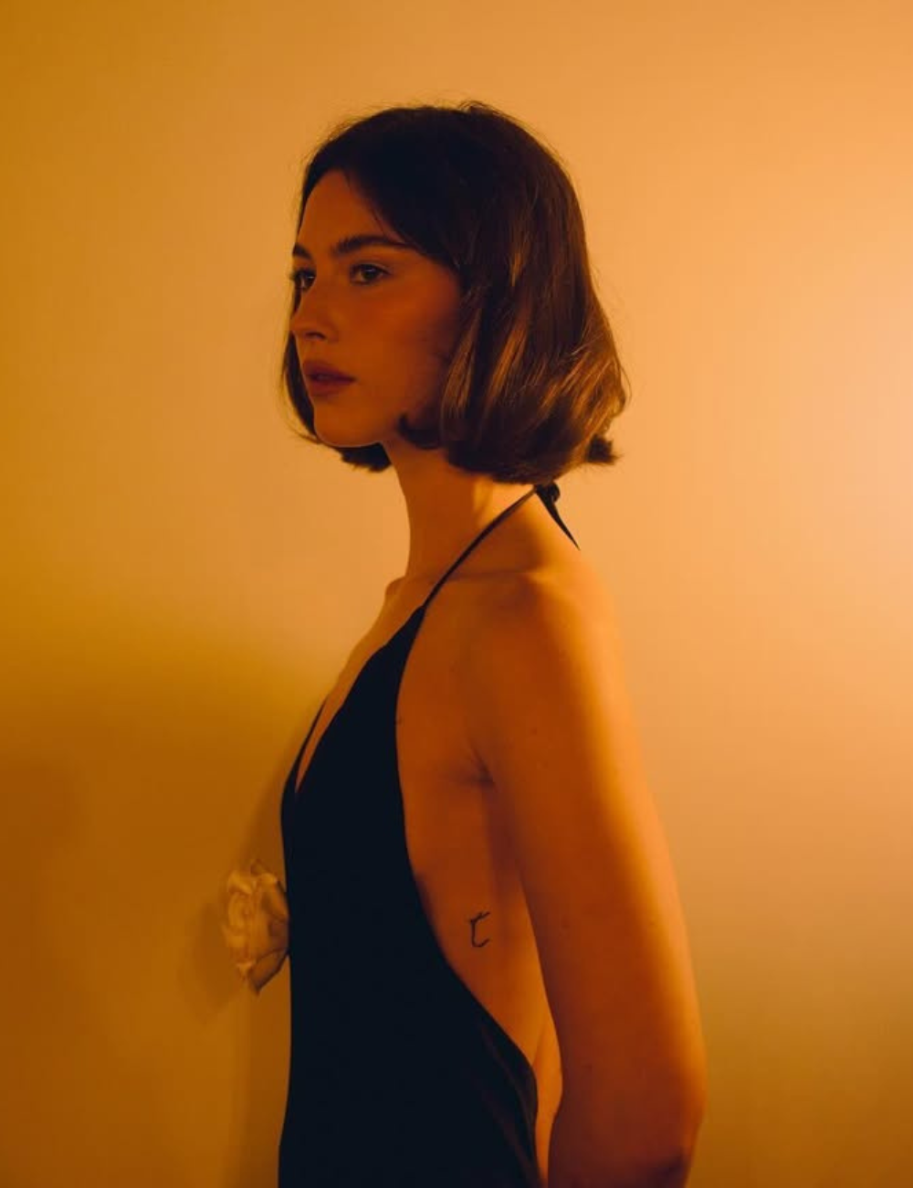 Modelo posa de perfil com long bob castanho e vestido preto de alças finas em luz âmbar, criando clima sofisticado.