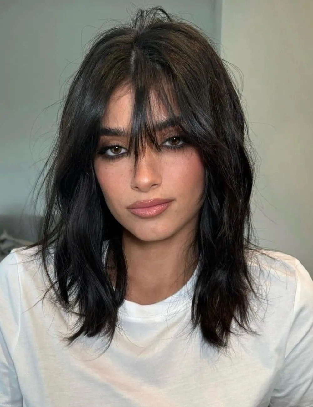 Modelo posa com long bob preto repicado, franja cortininha e camiseta branca, em clima sofisticado e suave.
