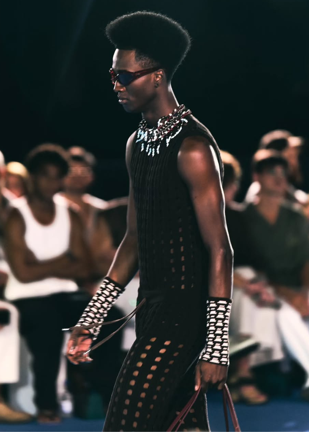 Desfile da MISCI na Sapucaí: modelo caminha com look preto vazado, mangas gráficas, colares e óculos, em clima sofisticado.