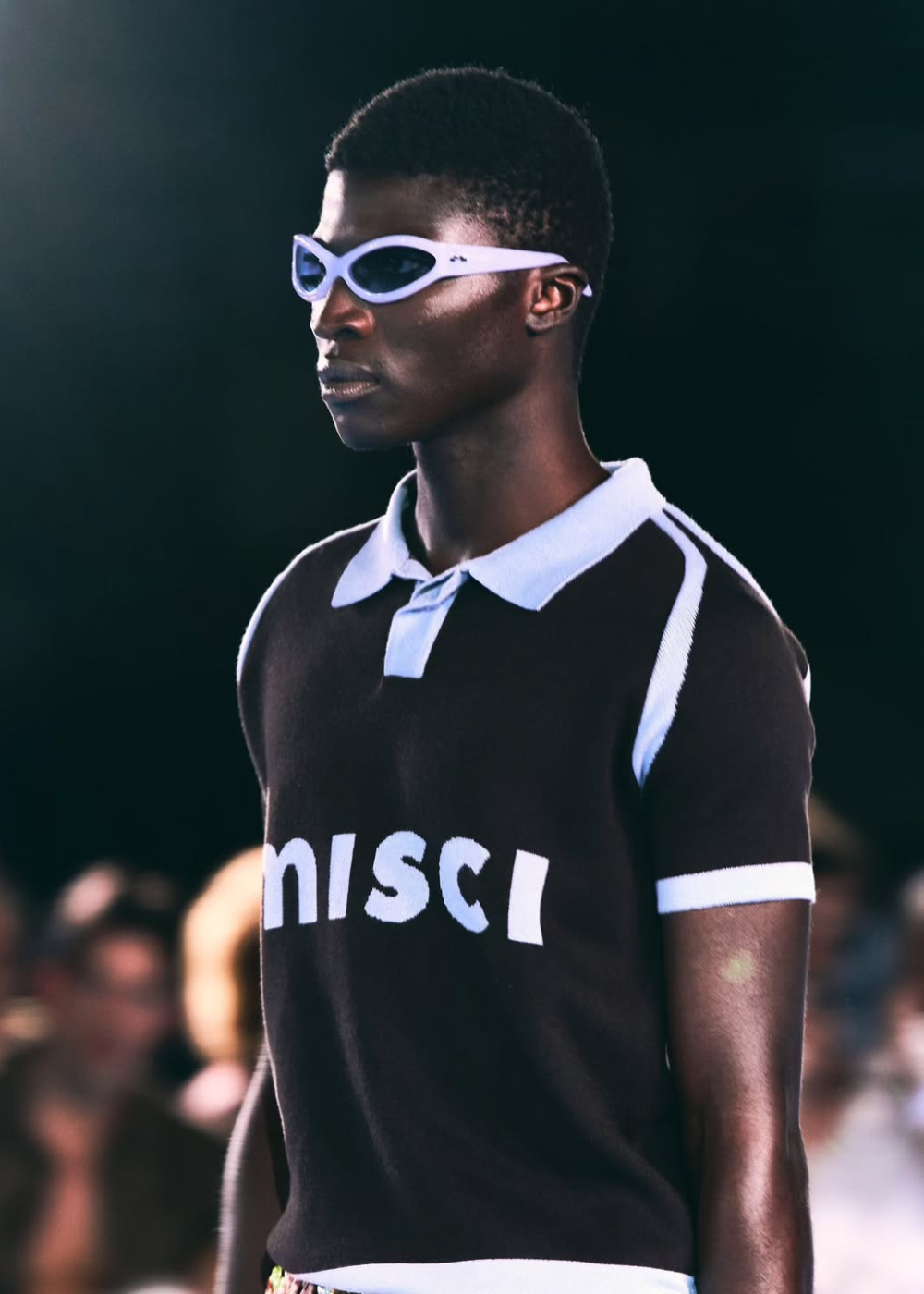 Desfile da MISCI na Sapucaí com modelo posa em polo preta de tricô e óculos lilás futuristas. Texto: misci