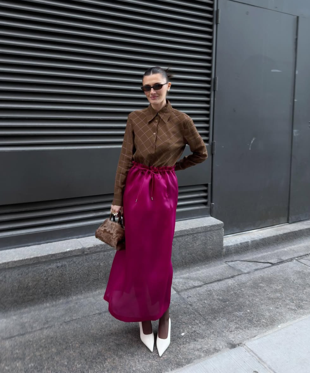 Looks com scarpin: modelo posa com camisa marrom xadrez, saia acetinada pink e bolsa texturizada em rua urbana sóbria.