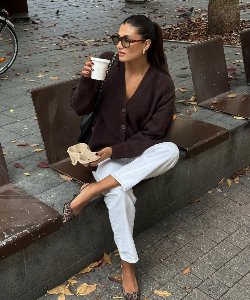 Looks com scarpin: modelo posa sentada com cardigan marrom, calça branca e scarpin animal print em clima urbano elegante.
