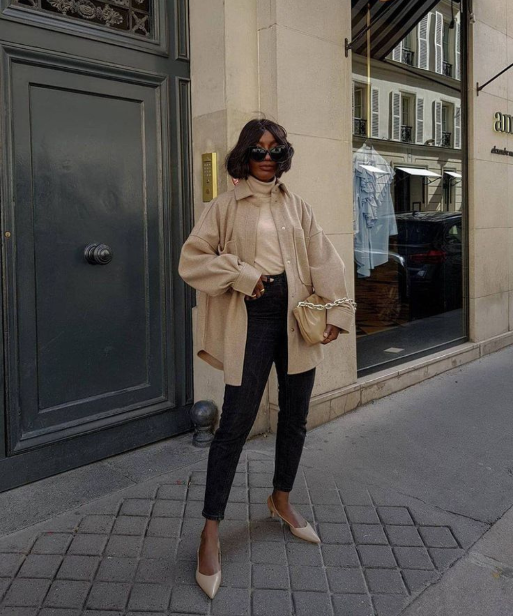 Looks com scarpin: modelo posa com camisa bege ampla, gola alta, jeans preto e bolsa neutra em rua elegante.