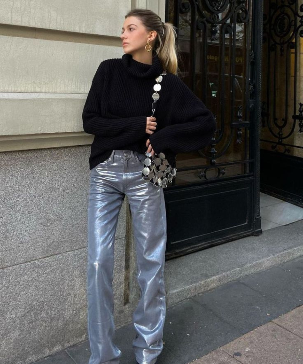 Modelo posa com calça resinada prata e tricô preto oversized, visual urbano sofisticado diante de fachada elegante.