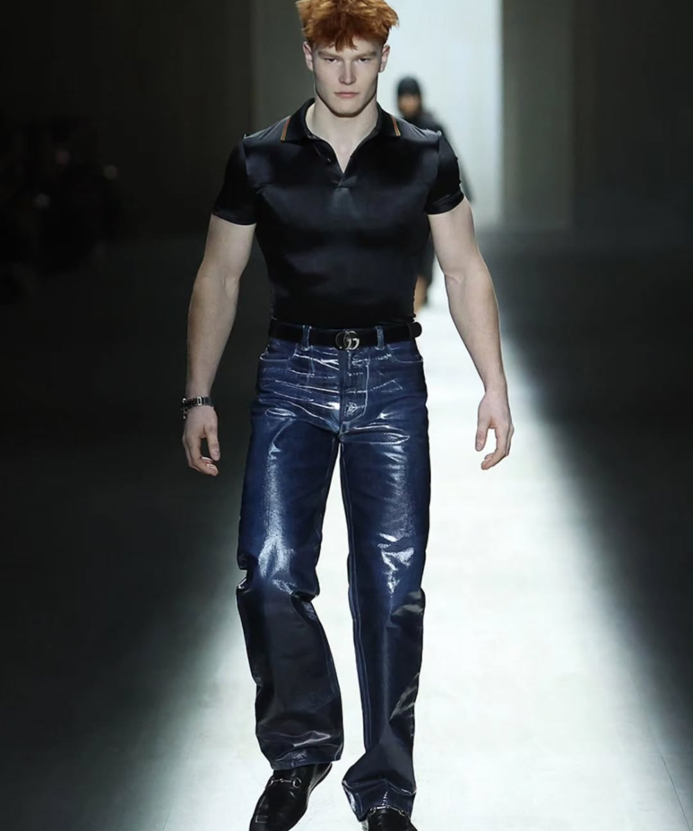 Modelo desfila com calça resinada azul de brilho intenso, polo preta ajustada e atitude sóbria na passarela escura.