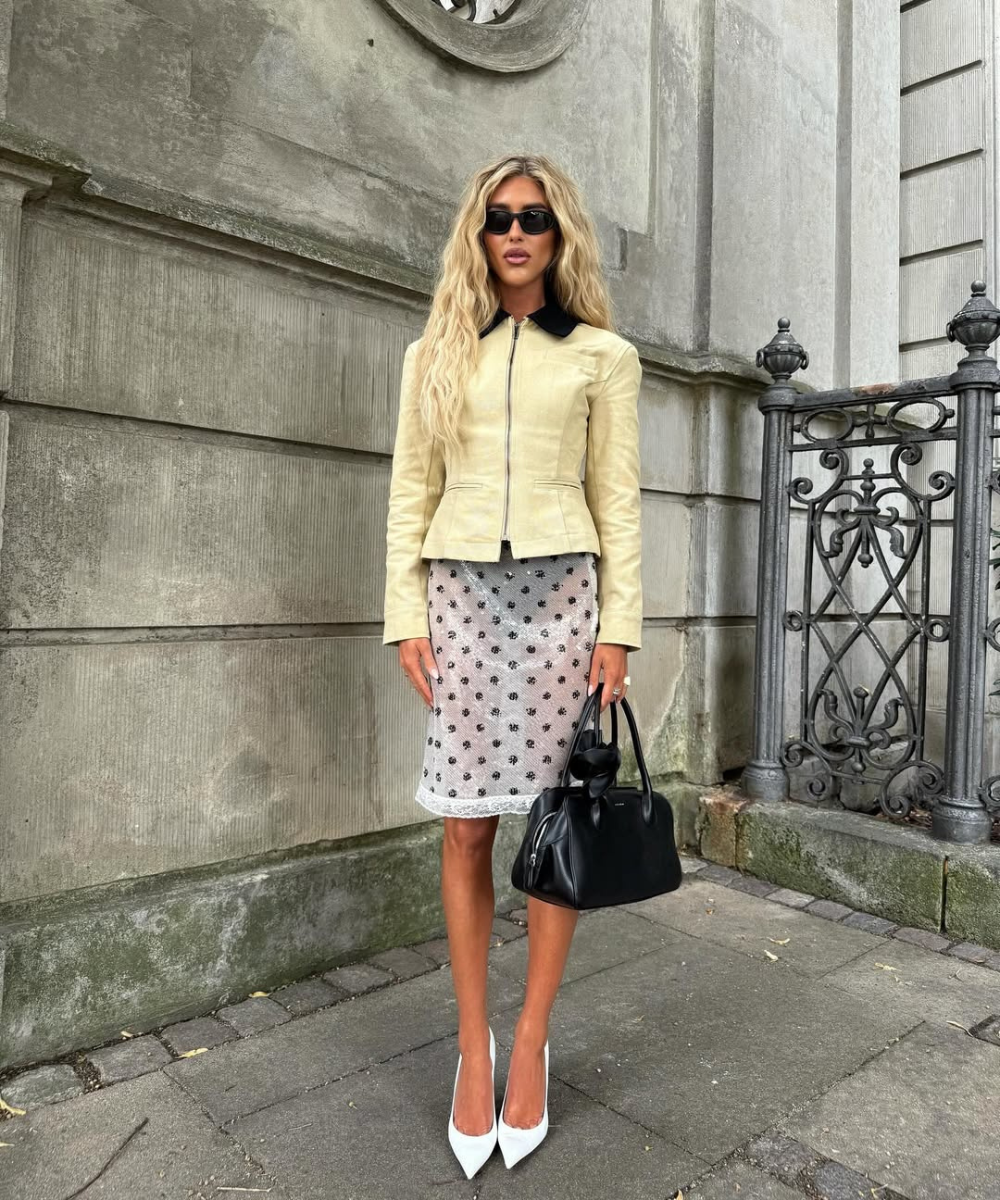 Looks com scarpin: modelo posa com jaqueta creme, saia floral rendada, bolsa preta e óculos em clima elegante.