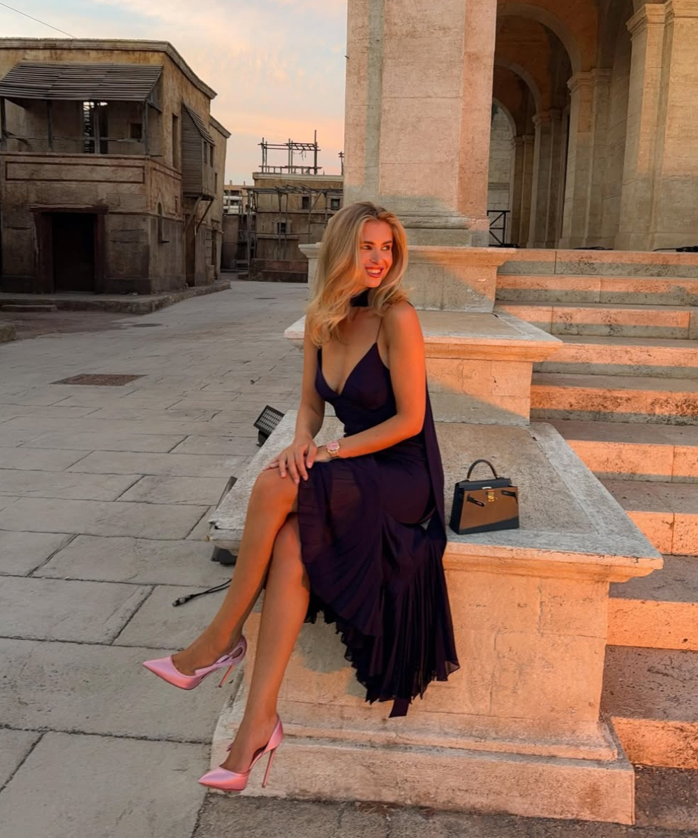 Looks com scarpin: modelo posa sentada em vestido roxo fluido e scarpin rosa, ao pôr do sol entre colunas de pedra.