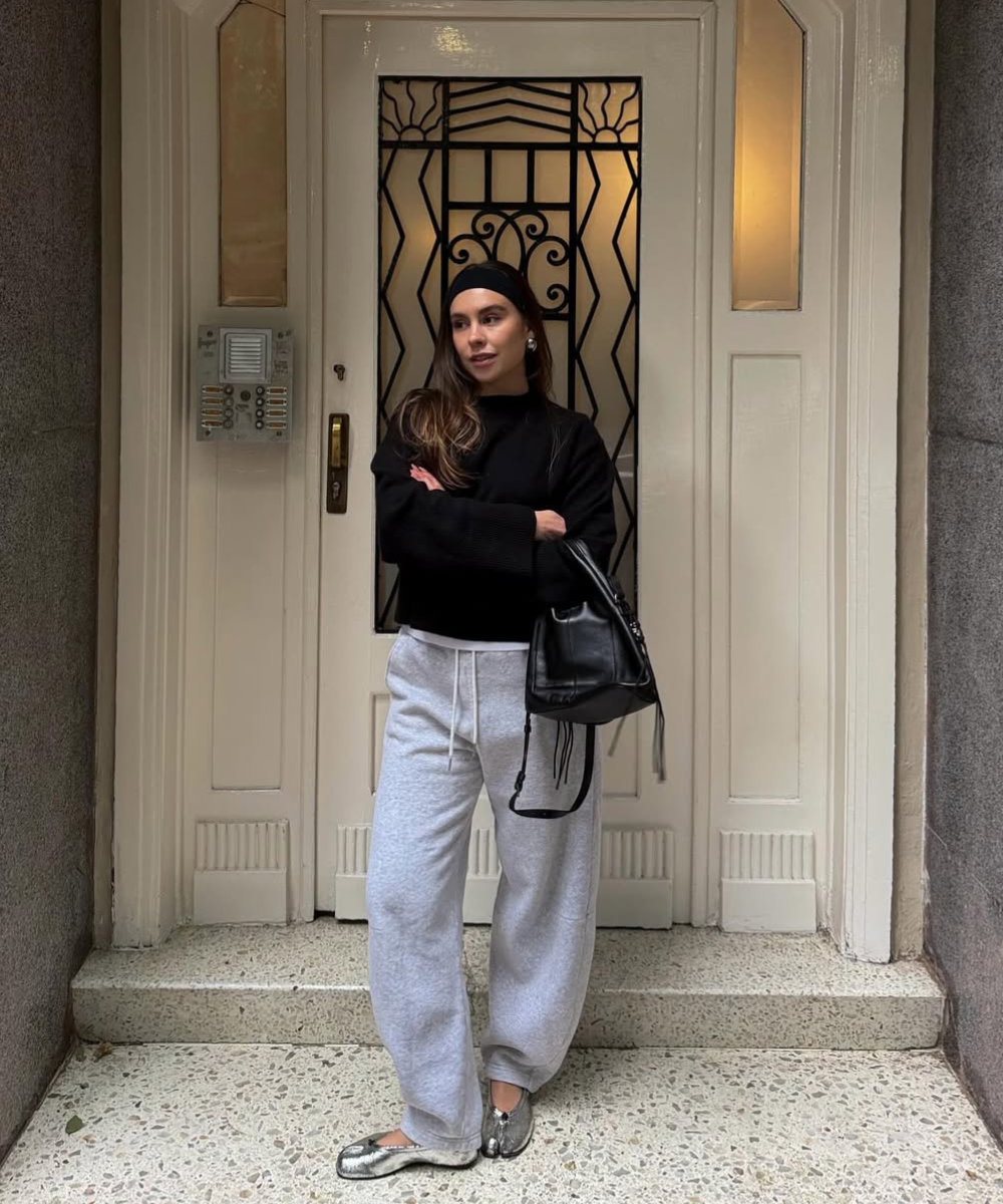 Looks com calça de moletom: modelo posa de braços cruzados com blusa preta, bolsa preta e cinza mescla em entrada elegante.