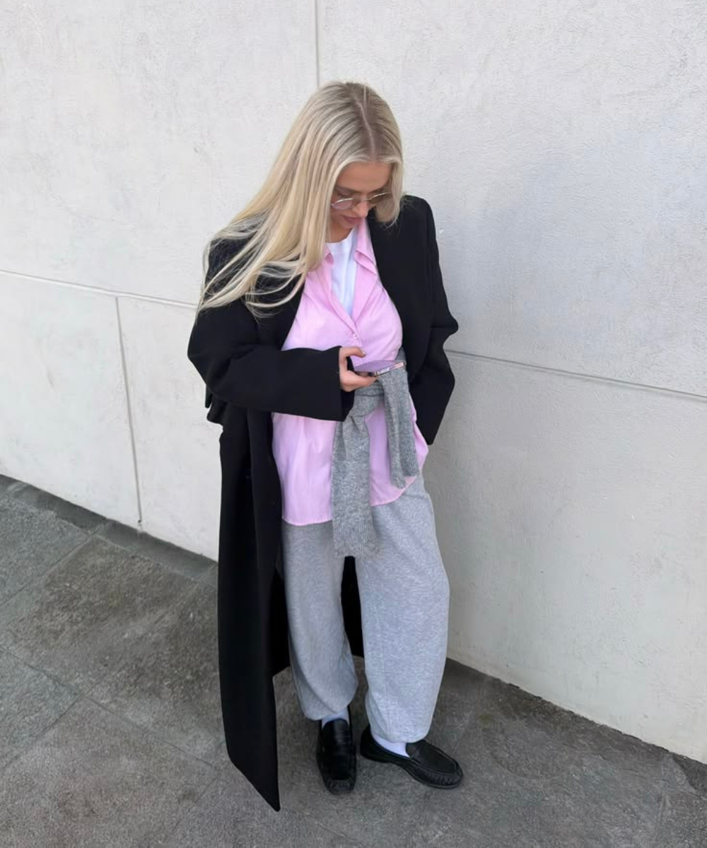 Looks com calça de moletom: modelo posa casual com camisa rosa, casaco preto oversized e cinza mescla urbano.