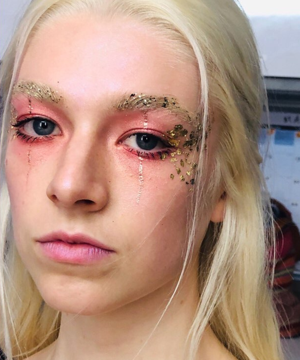 Modelo posa com maquiagens de euphoria em rosa e dourado, glitter nas sobrancelhas e lágrimas brilhantes, clima etéreo.