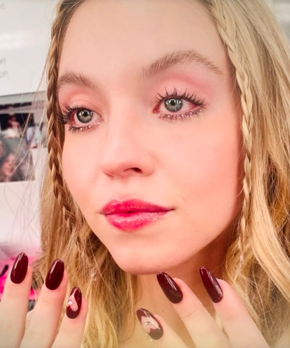 Maquiagens de euphoria em close com sombra rosa, gloss vibrante, tranças finas e unhas vinho em clima delicado e cool